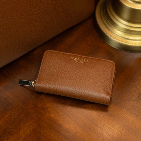 Von Baer Celeste small tan compact RFID leather zip-around wallet with coin pocket, displayed on a vintage table near an old lamp and a luxury tote bag.