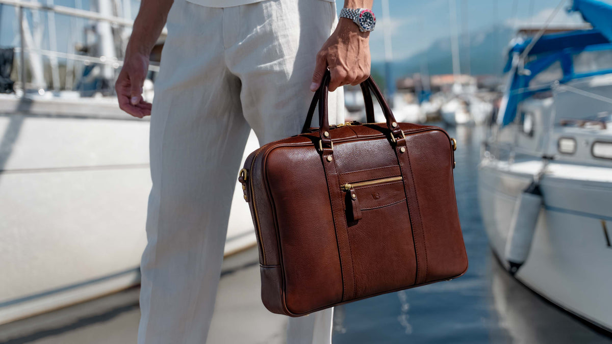 A man in white linen trousers with rolex watches carries a solid brown brown leather briefcase by Von Baer by a marina with boats in the background.