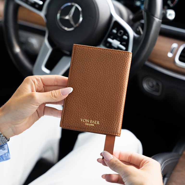 A woman holds a brown textured luxurious leather passport cover with a gold-tone Von Baer logo in a Mercedes car interior. The background shows a Mercedes-Benz steering wheel and dashboard.