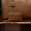 Von Baer Celeste small luxury compact tan leather zip-around wallet, displayed on a vintage wooden table next to a leather tote bag.