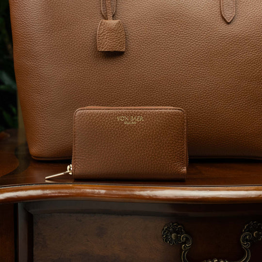 Von Baer Celeste small luxury compact tan leather zip-around wallet, displayed on a vintage wooden table next to a leather tote bag.