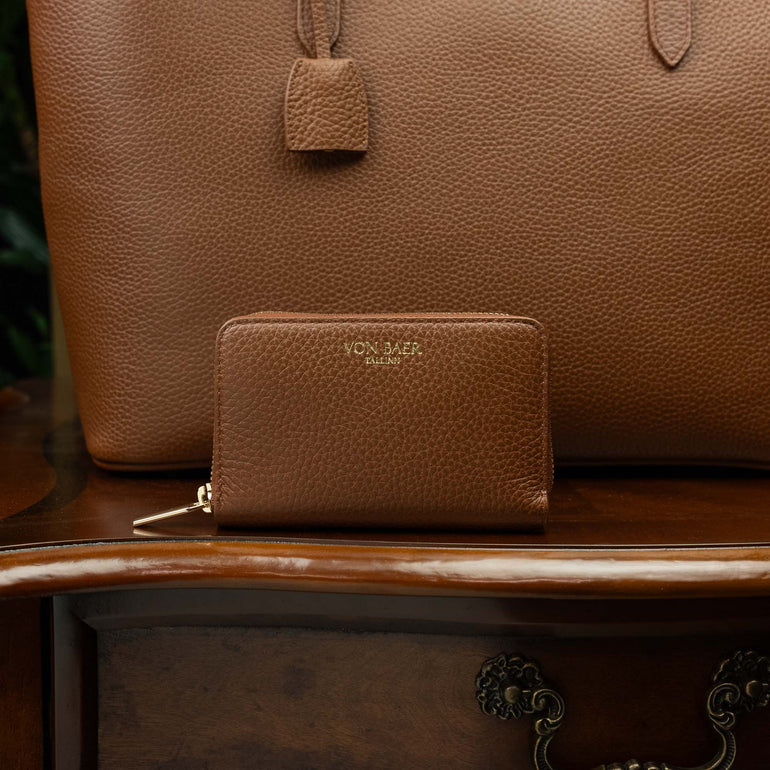 Von Baer Celeste small luxury compact tan leather zip-around wallet, displayed on a vintage wooden table next to a leather tote bag.