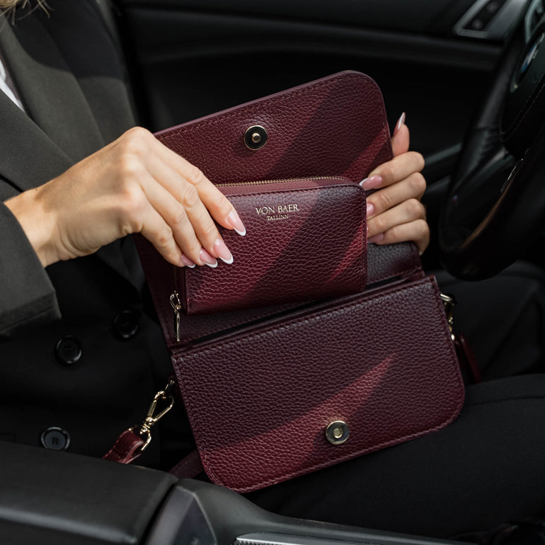 Von Baer Celeste small burgundy full-grain leather zip-around wallet demonstrated inside the Isabella small leather crossbody bag interior, held open inside a car setting with gold-tone hardware and gold-embossed Von Baer Tallinn branding.
