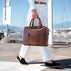 A woman in white trousers and black loafers carries a solid brown full grain leather briefcase by von baer with gold-tone hardware and a monogram logo, walking along a sunny outdoor harbor promenade.