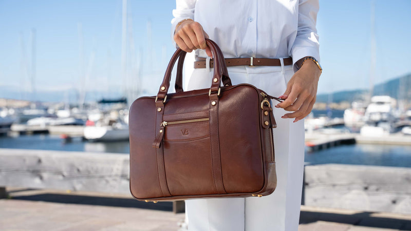A woman in a white shirt and trousers holds an elegant brown full grain leather briefcase-style bag by von baer with gold-tone hardware, standing by a marina with boats in the background.