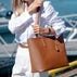 A woman in a white shirt and white pants carries a large tan pebbled leather tote bag by Von Baer with a matching shoulder strap in an outdoor harbor setting.