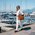 A beautiful woman walking along a marina boardwalk wearing a white shirt and white trousers in quit luxury style, carrying a large tan leather tote bag by von baer. Boats, mountains and water are visible in the background under a clear blue sky.