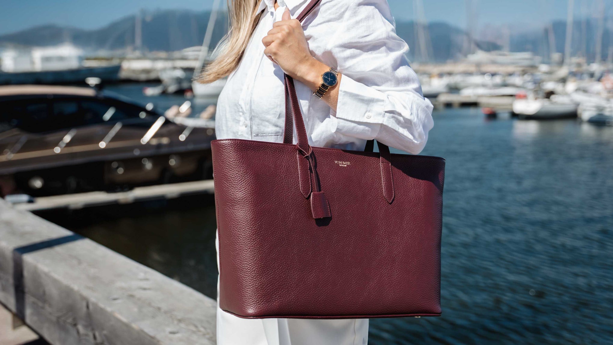 A woman in a white shirt carries a large burgundy leather tote bag by Von Baer with a tassel detail, set against a marina background with boats and water.