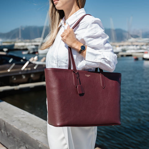 A woman in a white shirt and white pants in quiet luxury style carries a large burgundy pebbled leather tote bag by Von Baer with a front hanging clochette, standing by a sunny marina with water, mountains and boats in the background.