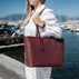 A woman in a white shirt and white pants in quiet luxury style carries a large burgundy pebbled leather tote bag by Von Baer with a front hanging clochette, standing by a sunny marina with water, mountains and boats in the background.