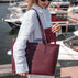 A woman at a marina carries a burgundy pebbled leather tote bag with matching wallet and matching elegant tote bag. The brand name 'Von Baer' is visible on the bag and wallet.