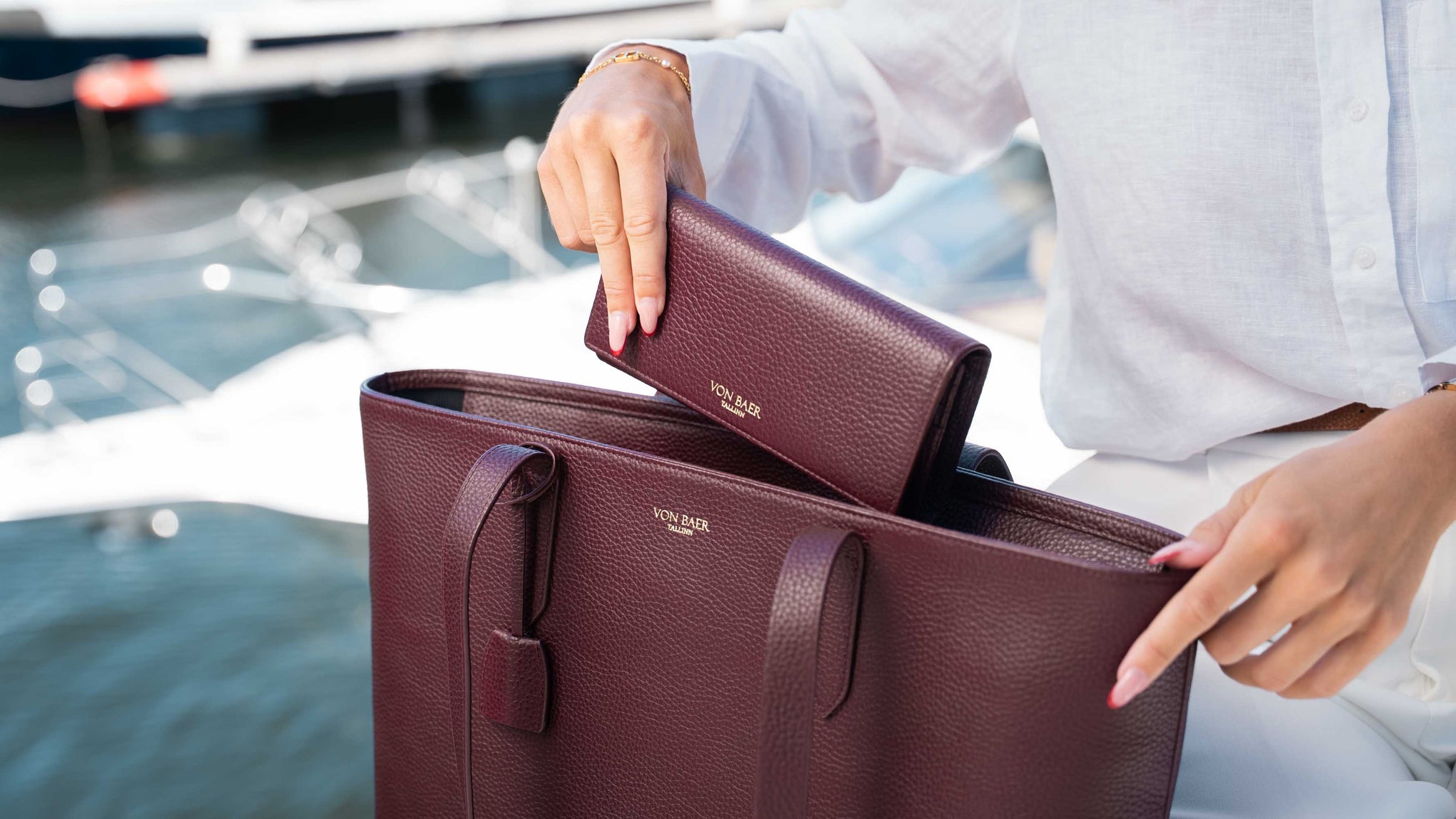 A person in a white shirt holds a matching burgundy leather tote bag and matching wallet with gold-tone Von Baer branding, set against a blurred outdoor background with water and boats.