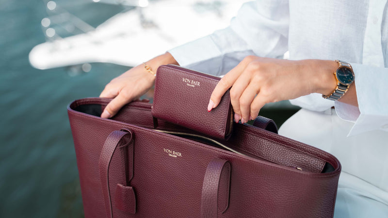 A person in a white outfit holds an open burgundy pebbled leather tote bag with a matching zip wallet, with water and a boat blurred in the background.