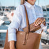 A woman in a white linen shirt carries a large brown pebbled leather tote bag with a matching zip-around wallet tucked into the bag's opening, set against a blurred marina background.