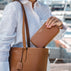 A person in a white linen shirt carries an amazing looking vb tan pebbled leather tote bag for women and holds a matching tan leather wallet. The background shows a blurred outdoor marina setting with boats.