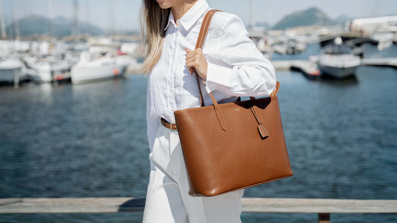 A woman in a white shirt and white pants carries a large high-quality cognac tan leather tote bag with a shoulder strap, standing by a marina with boats and water in the background in Italy.