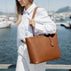 A woman in a white shirt and white pants in quiet luxury style carries a large cognac tan pebbled leather tote bag by Von Baer with tan shoulder straps, photographed outdoors near a marina with water, mountains and boats in the background.