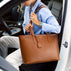 A woman in a light blue shirt and white pants sits in a car door holding a large vb brown pebbled leather tote bag with a small gold logo on the front.
