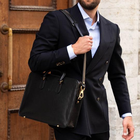 Man in a black suit holding a black leather bag against a stone wall and wooden door background