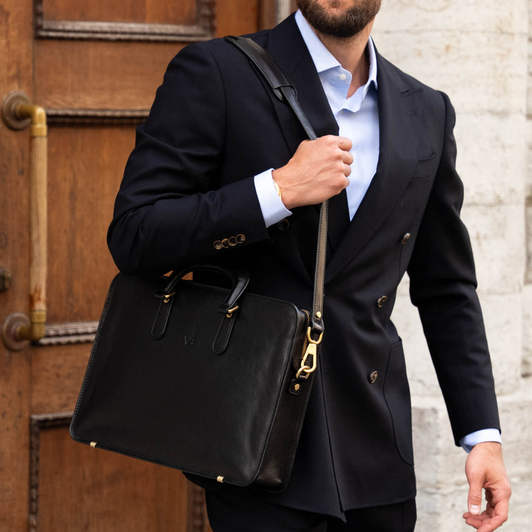 Man in a black suit holding a black leather bag against a stone wall and wooden door background