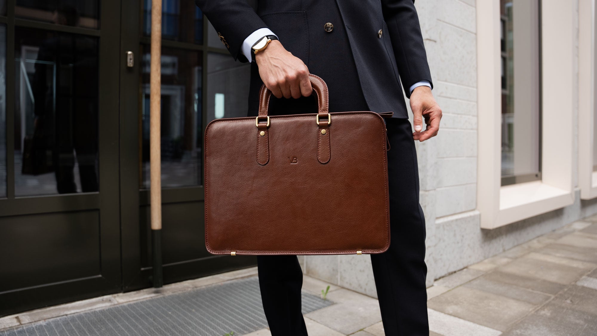 A businessman in an expensive navy blue suit holding a brown Von Baer elegant slim leather laptop bag.