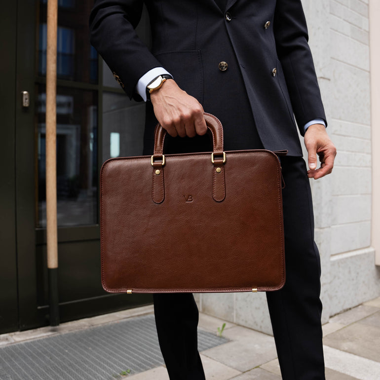 Person in navy blue suit holding a luxury brown leather briefcase outdoors