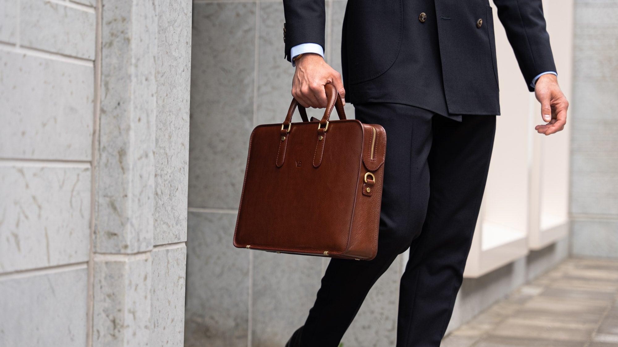 A businessman in a navy blue suit holding a brown Von Baer elegant slim leather laptop bag near a bank.
