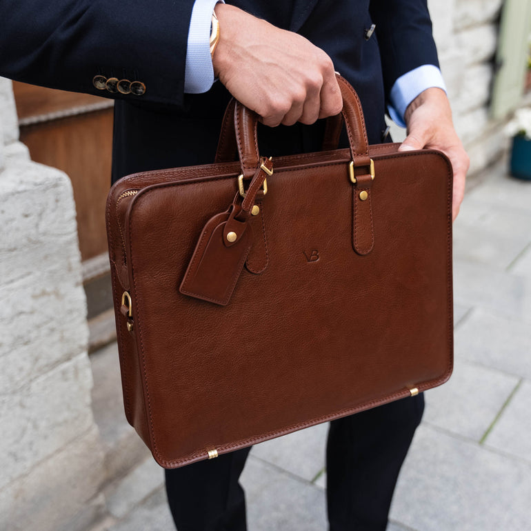 A businessman holding a brown Von Baer elegant slim leather laptop bag with a luggage tag.