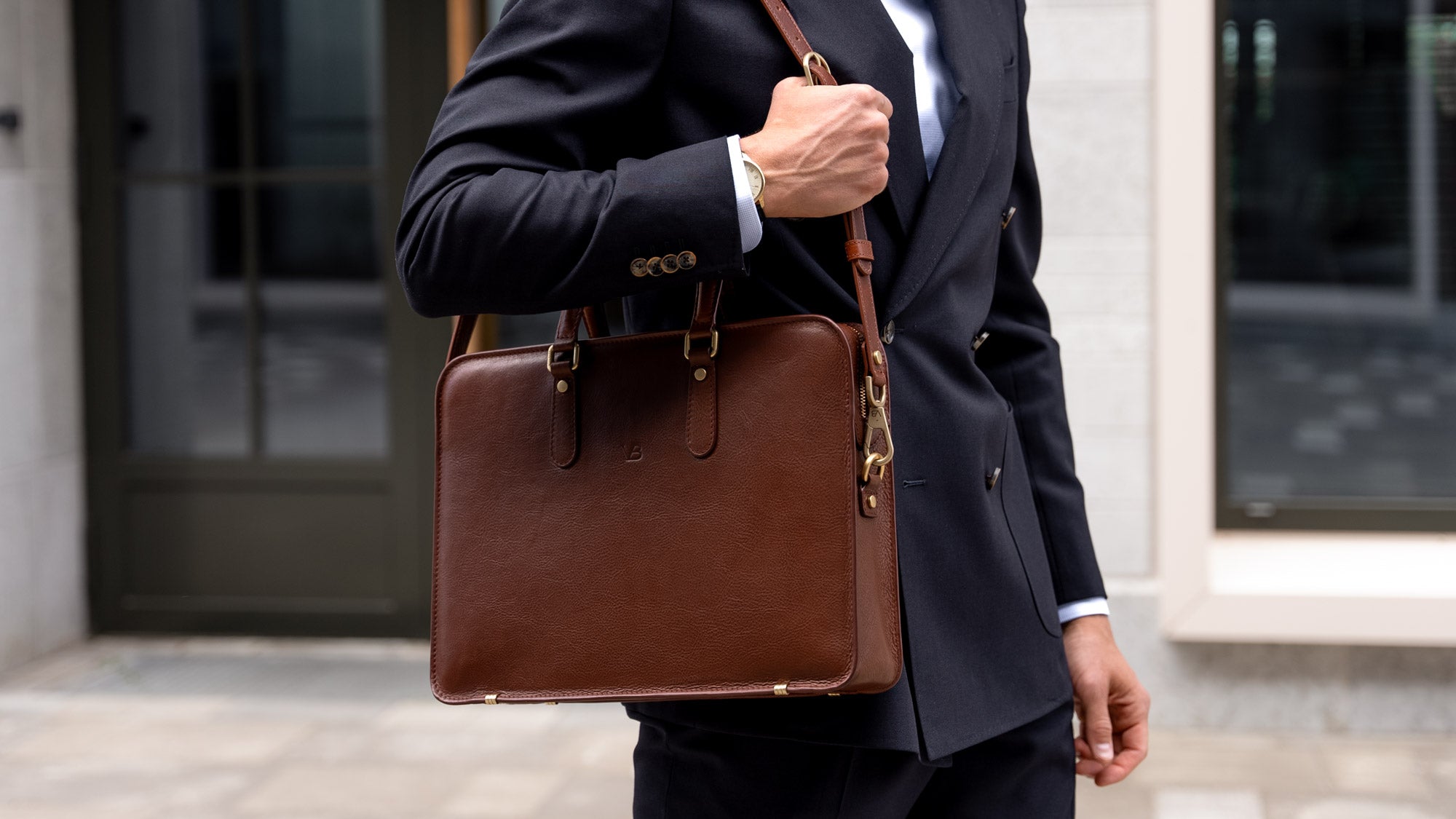 An elegant Von Baer slim leather laptop bag with a shoulder strap, for documents and essentials, held by a businessman in a classic navy blue suit.
