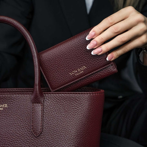 Von Baer Elise trifold wallet and Estelle small tote bag in burgundy pebbled leather held together in a luxury car setting, showing gold foil brand lettering.
