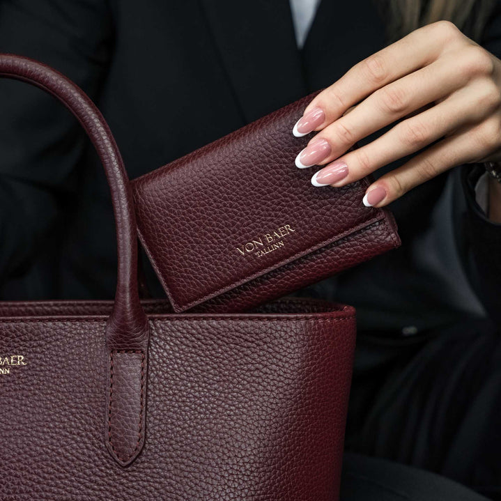 Von Baer Elise trifold wallet and Estelle small tote bag in burgundy pebbled leather held together in a luxury car setting, showing gold foil brand lettering.

