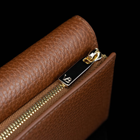 Von Baer Elise women's cognac pebbled leather trifold wallet shown in a close-up detail shot on a solid black background, featuring gold-tone zipper hardware and a monogram zipper pull.