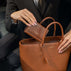 Von Baer Elise trifold wallet and Estelle small tote bag in cognac pebbled leather held by a person in a dark blazer inside a luxury car, showing the wallet being placed into the bag.