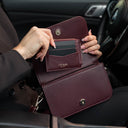 Von Baer Isabella small burgundy pebbled leather crossbody bag shown with the Essence women's card case fitting inside, held in a car interior and featuring embossed Von Baer Tallinn branding.