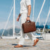 A man in a white shirt and trousers walks along a cobblestone harbor path carrying a solid brown full grain leather briefcase, with boats and blue sky in the background.