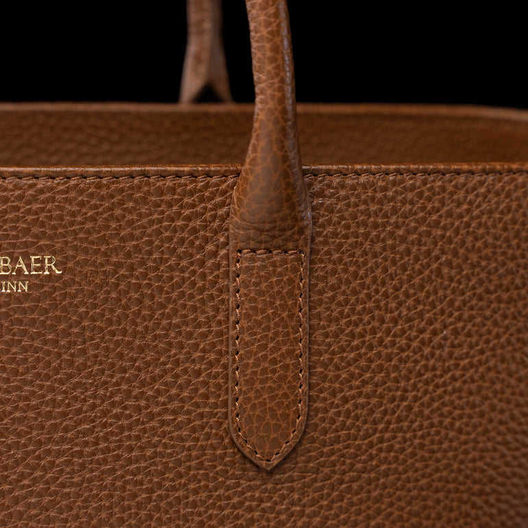 Close-up of a brown pebbled leather handbag with a top handle, showing a stitched tab detail and partial gold-toned 'VON BAER TALLINN' branding on the left side against a black background.
