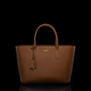 A brown pebbled leather tote bag with double top handles, a hanging leather tag, and a small gold-tone logo plaque on the front, photographed against a solid black background.