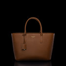 A brown pebbled leather tote bag with double top handles, a hanging leather tag, and a small gold-tone logo plaque on the front, photographed against a solid black background.