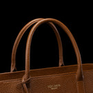 Close-up of a brown pebbled leather handbag with double top handles and a small gold-toned VON BAER logo on the front, set against a solid black background.