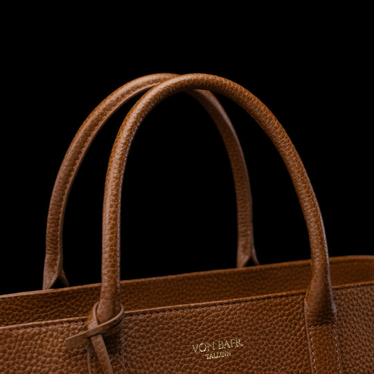 Close-up of a brown pebbled leather handbag with double top handles and a small gold-toned VON BAER logo on the front, set against a solid black background.