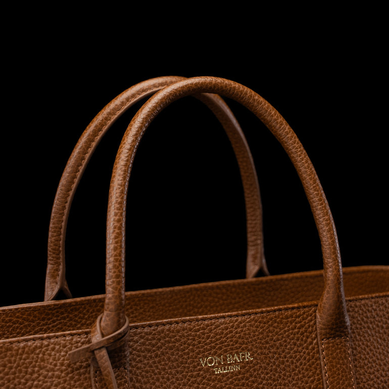 Close-up of a brown pebbled leather handbag with double top handles and a small gold-toned VON BAER logo on the front, set against a solid black background.