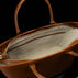 Close-up of the interior of a brown pebbled leather tote bag, showing a cream-colored fabric lining, a zippered compartment, and a side pockets with gold-tone hardware against a black background.