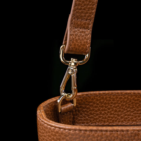 Close-up of a brown pebbled leather bag strap attached with a gold-tone metal clasp against a black background.
