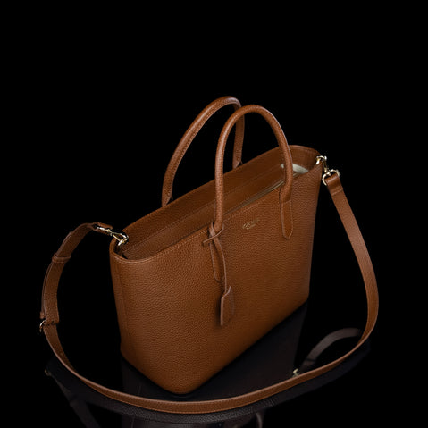 A brown pebbled leather handbag with double top handles and a detachable shoulder strap, photographed on a reflective black surface against a solid black background.