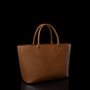 A structured tan leather tote bag with a pebbled texture and double top handles, photographed against a solid black background with a subtle reflection below.