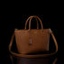A brown pebbled leather handbag with dual top handles, a matching shoulder strap, and a small hanging leather tassel, photographed against a black background with a reflective surface below.