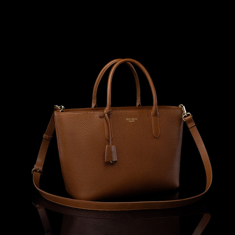 A brown pebbled leather handbag with dual top handles, a matching shoulder strap, and a small hanging leather tassel, photographed against a black background with a reflective surface below.