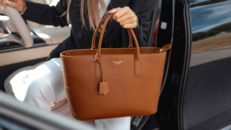 A woman in a black blazer and white pants sits in a car door holding a brown pebbled leather tote bag with gold-tone hardware and the brand name 'VON BAER' visible on the front.