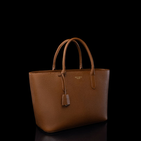 A brown pebbled Estelle leather tote bag with double top handles and a hanging leather tag, photographed against a solid black background.