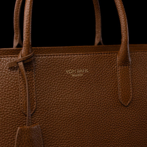 Close-up of a brown pebbled leather tote bag with gold-toned 'VON BAER' branding on the front, set against a solid black background.